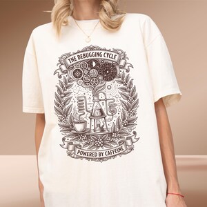 May include: An off-white short-sleeve t-shirt featuring a dark brown graphic print. The design depicts a laboratory setup with a coffee cup, gears, and botanical elements, with the text THE DEBUGGING CYCLE and POWERED BY CAFFEINE.