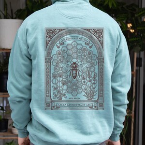 May include: A light blue hooded sweatshirt featuring a dark brown graphic on the back. The design depicts a central bee within a honeycomb pattern, surrounded by botanical illustrations and an architectural arch with the text Apis Regina Et Natura.