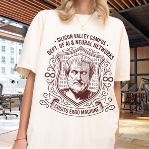 Op de afbeelding: Een off-white t-shirt met korte mouwen en een donkerbruine afbeelding van een filosoof met een print van een printplaat op de helft van het gezicht, in een schild met binaire code en de tekst SILICON VALLEY CAMPUS DEPT. OF AI & NEURAL NETWORKS en COGITO ERGO MACHINE.
