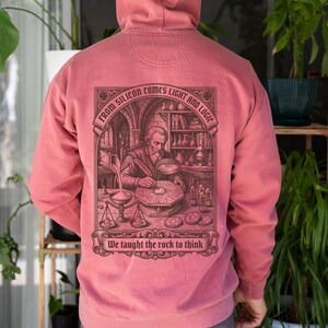Puede incluir: Una sudadera con capucha de color rosa empolvado con un gráfico marrón oscuro en la espalda. La ilustración muestra a una figura barbuda trabajando en un escritorio con herramientas científicas y el texto: FROM SILICON COMES LIGHT AND LOGIC y We taught the rock to think.
