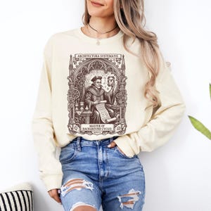 Puede incluir: Una sudadera de manga larga color crema con un estampado gráfico marrón oscuro de un erudito sosteniendo un reloj de arena y un pergamino. El diseño incluye arcos arquitectónicos intrincados, engranajes y el texto ARCHITECTURA SYSTEMATIS y MASTER OF BACKGROUND CYCLES MDXVII.
