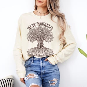 May include: A cream-coloured long-sleeve sweatshirt featuring a dark brown graphic print of a stylised tree with intricate circuit board roots and branches. The text "RETE NEURALIS" is arched above the tree, and "CONEXIO ETHERIS" is on a banner below.