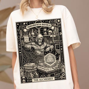 Könnte beinhalten: Ein cremefarbenes T-Shirt mit einem schwarzen Grafikdruck eines kapuzenbekleideten Alchemisten, der an einem Schreibtisch mit digitalen Elementen arbeitet. Das Design enthält den Text "TRANSMUTATIO DIGITALIS" und "THE ALCHEMIST" sowie Symbole, Binärcode und eine leuchtende geometrische Form.