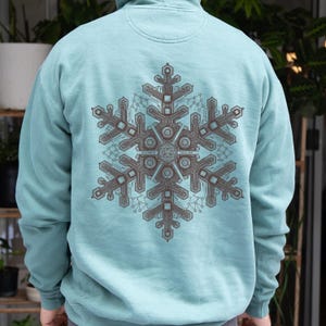 Puede incluir: Una sudadera con capucha de color azul claro que presenta un gráfico grande e intrincado en la espalda. El diseño es un copo de nieve estilizado compuesto por patrones de circuitos en marrón oscuro, líneas geométricas y textos.