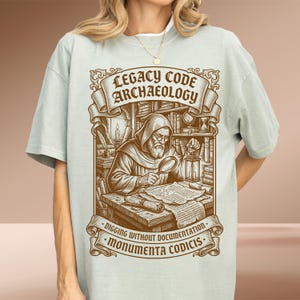 Puede incluir: Una camiseta de color verde salvia claro con un estampado gráfico marrón de una figura encapuchada examinando un documento con una lupa y una linterna. El texto dice LEGACY CODE ARCHAEOLOGY, DIGGING WITHOUT DOCUMENTATION y MONUMENTA CODICIS.