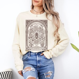 May include: A cream-colored long-sleeve sweatshirt featuring a brown graphic print of a bee centered within a hexagonal honeycomb pattern, surrounded by floral illustrations and an architectural archway with the text APIS REGINA ET NATURA.