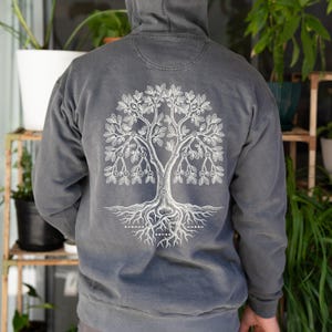May include: A charcoal gray hooded sweatshirt featuring a white graphic on the back. The design depicts a detailed oak tree with intricate roots and branches, incorporating small circles containing numbers and Morse code symbols at the base.