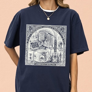 Puede incluir: Una camiseta azul marino con un estampado gráfico blanco de una escena de laboratorio alquímico con un aparato de destilación, velas y libros. El texto dice "TRANSFORMATIO CAFFEINAE IN BINARIA" y "ALEMBICUM ET DISTILLATIO" en un borde.