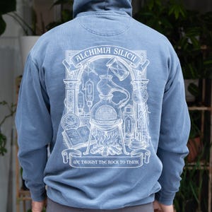 May include: A dusty blue hooded sweatshirt featuring a white graphic print on the back. The design depicts an alchemical laboratory scene with a central flask over a fire, an hourglass, a book, and the text ALCHIMIA SILICII and WE TAUGHT THE ROCK TO THINK.