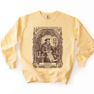 May include: A pale yellow crewneck sweatshirt featuring a dark brown graphic print of a scholar holding an hourglass and a scroll. The design includes intricate architectural arches, gears, and the text ARCHITECTURA SYSTEMATIS and MASTER OF BACKGROUND CYCLES.