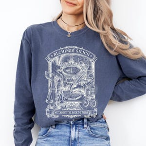 May include: A blue long-sleeve shirt featuring a white graphic print of an alchemical laboratory scene with a hand using a quill on a circuit board. The text reads ALCHIMIA SILICII at the top and WE TAUGHT THE ROCK TO THINK at the bottom.