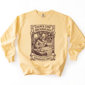 Op de afbeelding: Een lichtgeel sweatshirt met ronde hals en een donkerbruine grafische print van een figuur met een kap die een document onderzoekt met een vergrootglas. De tekst luidt LEGACY CODE ARCHAEOLOGY, DIGGING WITHOUT DOCUMENTATION en MONUMENTA CODICIS.