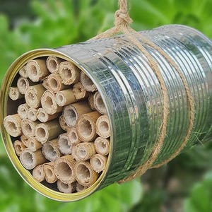 Op de afbeelding: Een cilindervormig metalen blik gevuld met talloze holle bamboerietjes van verschillende diameters. De zilverkleurige houder is omwikkeld met natuurlijk bruin touw, dat aan de bovenkant een lus vormt om het geheel op te kunnen hangen.