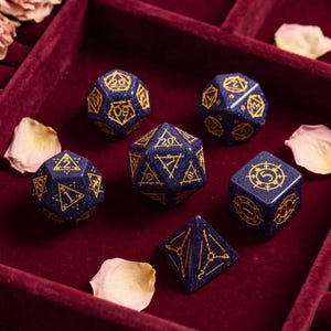 May include: A set of six polyhedral dice made from dark blue glittery material with gold engraved numbers and geometric symbols. The collection includes a d20, d12, d10, d8, d6, and d4, each featuring intricate line work and clear numerical markings.