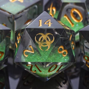 May include: A twenty-sided die featuring a translucent dark grey resin shell with a vibrant green glitter core. The die displays gold-coloured numbers and a gold biohazard symbol on the top face, with other numbers visible on the surrounding facets.
