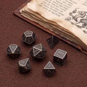 Op de afbeelding: Een set van zeven veelzijdige metalen dobbelstenen met een antieke bronzen afwerking en zwart gegraveerde cijfers. De collectie bevat een d20, d12, d10, d8, d6, d4 en een procentuele dobbelsteen, allemaal met scherpe randen en een metaalglans, bij een boek.