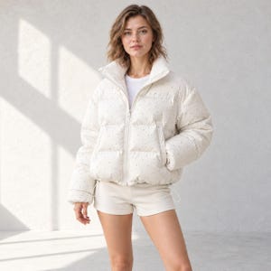 May include: A white puffer jacket featuring a subtle black speckled pattern, a high collar, and a full-length front zipper. The jacket has long sleeves with elastic cuffs and two side pockets, worn over a plain white t-shirt and white shorts.
