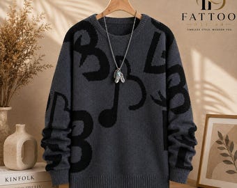 May include: A charcoal grey long-sleeve knit jumper featuring a large black abstract pattern of musical notes and numbers. The jumper has a crew neckline and ribbed cuffs and hem. A silver-toned necklace with a feather pendant hangs from the collar.