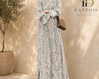 May include: A floor-length white dress featuring a black speckled pattern, long sleeves with flared cuffs, and a side slit. The garment is paired with a tan handbag and open-toe sandals. The text FATTOO DESIGN TIMELESS STYLE MODERN YOU is visible.