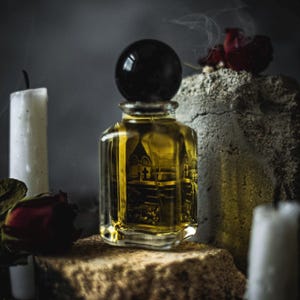 May include: A square glass perfume bottle filled with a golden liquid, featuring a black spherical cap and a label depicting a dark architectural scene. The bottle is surrounded by white candles and dark red roses in a dimly lit, atmospheric setting.
