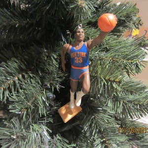 May include: A Christmas tree ornament featuring a basketball player wearing a blue and orange jersey with the text "New York 33". The player is holding a basketball and is in a mid-air jump shot pose.