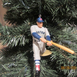 May include: A baseball player ornament wearing a white and blue uniform with the word "BOSTON" on the front. The player is holding a wooden bat and is wearing a blue cap with a red logo.