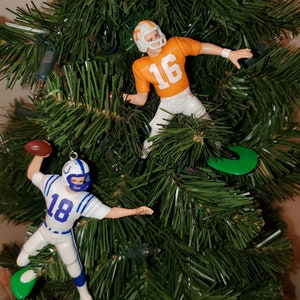 May include: Two football player ornaments hanging on a green artificial Christmas tree. The player on the left is wearing a white jersey with blue trim and the number 18. The player on the right is wearing an orange jersey with the number 16.