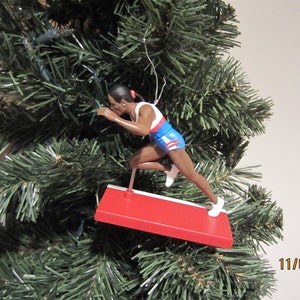 May include: A Christmas ornament featuring a figure of a runner in a blue and red uniform, with a white stripe on the side, running over a red and white hurdle. The ornament is hanging on a green Christmas tree.