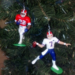 May include: Two football player ornaments hanging on a green Christmas tree. The player on the left is wearing a red and white uniform with the number 22. The player on the right is wearing a blue and white uniform with the number 7.