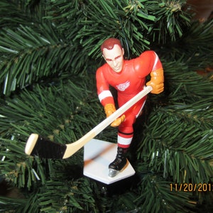May include: A red and white hockey player ornament with a stick and a white base. The player is wearing a red jersey with a white wing logo and white pants. The ornament is on a green Christmas tree.