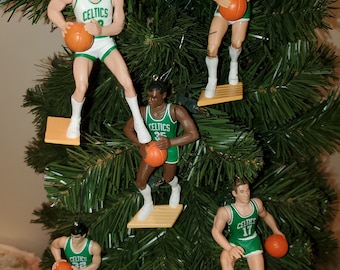 May include: A collection of basketball player ornaments, each depicting a Boston Celtics player in a green and white uniform, holding an orange basketball. The ornaments are hanging on a green Christmas tree. Each player is standing on a small, rectangular platform.