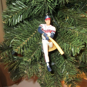 May include: A baseball player ornament wearing a red and white uniform with a baseball bat in hand. The ornament is hanging from a green artificial Christmas tree.