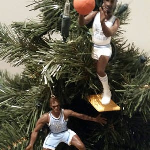 May include: Two basketball player ornaments hanging on a green artificial Christmas tree. The ornaments are of a basketball player wearing a blue and white uniform with the text "North Carolina" on the front. One ornament is holding a basketball.