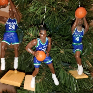 May include: Three Dallas Mavericks basketball player ornaments, each wearing a blue and green uniform with a basketball. The ornaments are hanging on a green Christmas tree.