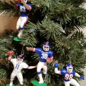 May include: Four blue and white football player ornaments hanging on a green Christmas tree. The players are wearing helmets and jerseys with the numbers 88, 30, 11, and 98.