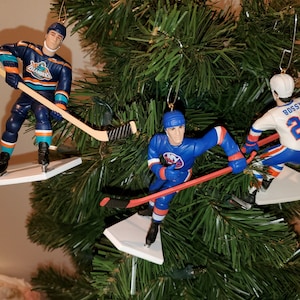 May include: Three hockey player Christmas ornaments, each with a white base and a hockey stick. The player on the left is wearing a blue and orange jersey with the word "Islanders" on it. The player in the middle is wearing a blue and orange jersey with the letters "NY" on it. The player on the right is wearing a white jersey with the name "Bossy" and the number "22" on it.