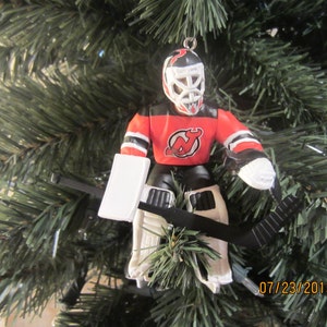 May include: A red, white, and black hockey goalie ornament with a New Jersey Devils logo on the front. The ornament is hanging on a green Christmas tree.