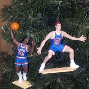 May include: Two blue and white basketball player ornaments hanging from a green Christmas tree. The player on the left is holding a basketball and is wearing number 43. The player on the right is wearing number 25 and is standing on a wooden platform.