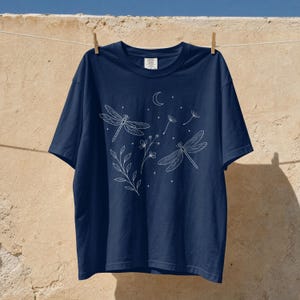 Op de afbeelding: Een marineblauw t-shirt met korte mouwen en een witte lijntekening van twee libellen, een maansikkel, kleine sterren, een takje met bladeren, een klein bloemtakje en paardenbloemzaadjes. Het shirt heeft een ronde hals.