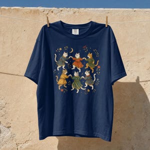 Può includere: Una t-shirt blu navy a maniche corte con una grafica centrale di sei gatti che indossano tuniche e si tengono per le zampe in cerchio. L'illustrazione include piccoli elementi floreali, una luna crescente e stelle attorno ai gatti.