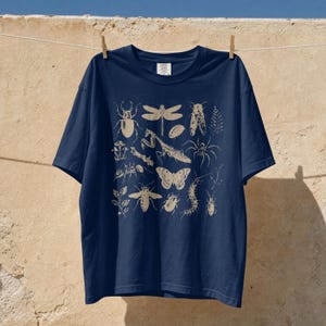 Op de afbeelding: Een marineblauw t-shirt met korte mouwen en een beige grafische print van diverse insecten, waaronder een libel, een bidsprinkhaan, een vlinder, een kever en een spin, gerangschikt in een rasterpatroon in het midden van de borst.