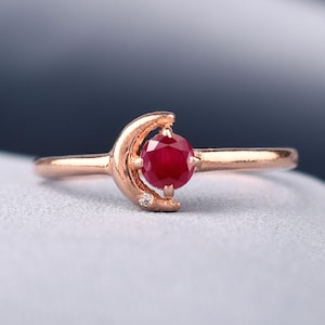 Könnte beinhalten: Ein Ring aus Roségold mit einer dünnen Ringschiene, einem Halbmond-Motiv und einem runden, facettierten, tiefroten Rubin in der Mitte. Ein winziger, klarer, rund geschliffener Akzentstein ist an der unteren Spitze des Halbmonds eingelassen.
