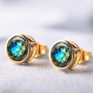 May include: A pair of round stud earrings featuring polished gold-tone settings and cabochon-cut labradorite gemstones. The stones display a vibrant blue and yellow iridescent flash with natural internal inclusions visible within the surface.