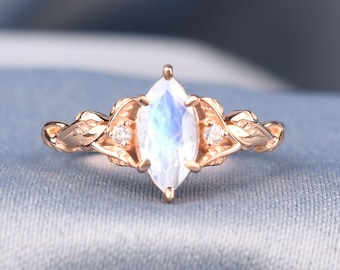 May include: A rose gold ring featuring a marquise-cut moonstone centre stone flanked by two small round clear accent stones. The band is designed with a detailed leaf motif that extends from the central setting along both sides of the ring.