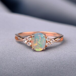 May include: A rose gold ring featuring a central oval-shaped opal gemstone with a play of color. The opal is flanked on each side by three small, round, clear, faceted stones set in a prong setting. The band has a split design near the setting.
