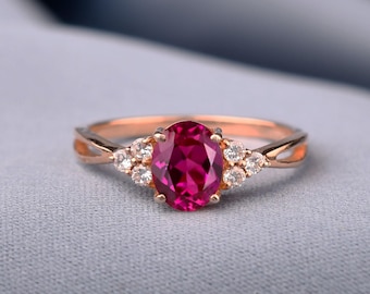 May include: A rose gold ring featuring a central oval-cut vibrant pink gemstone flanked by three small round-cut clear stones on each side. The band has a split design that tapers towards the back, showcasing a polished metallic finish.