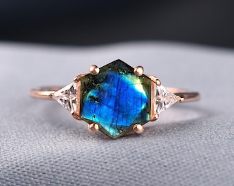May include: A rose gold ring featuring a central hexagonal labradorite gemstone with a vibrant blue and green iridescent flash, flanked by two small clear triangular stones on either side, all set in a delicate metal band with six prongs.