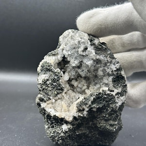 Könnte beinhalten: Ein raues, dunkelgraues Vulkangestein mit einer zentralen Aushöhlung, die mit kleinen, durchscheinenden, klaren Kristallstrukturen gefüllt ist. Die Außenseite des Steins hat eine poröse, texturierte Oberfläche, die einen Kontrast zu den reflektierenden, gezackten Innenkristallen bildet.