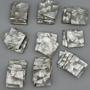 Könnte beinhalten: Eine Sammlung quadratischer Fliesen, die in neun separaten Stapeln angeordnet sind. Jede Fliese weist ein Mosaikmuster aus irisierenden, weißen und grauen Perlmuttfragmenten mit einer glatten, reflektierenden Oberfläche und variierten geometrischen Formen auf.