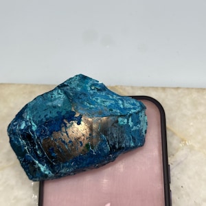 May include: A raw, irregularly shaped mineral specimen featuring vibrant shades of blue and teal with a prominent, smooth, metallic bronze-colored patch on its surface, resting atop a pink smartphone with a visible white Apple logo on the back.